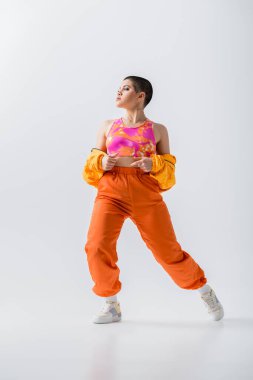 modern individual, warm clothing, fashionable young woman with short hair posing in puffer jacket and vibrant pants on grey background, outerwear, modern subculture, youthful energy, full length 