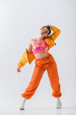 personal style, fashionable young woman with short hair posing in puffer jacket and orange pants on grey background, outerwear, modern subculture, youthful energy, full length 