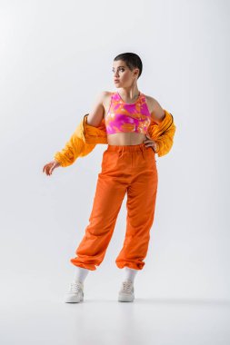 fashion trend, warm clothing, fashionable young woman with short hair posing with hand on hip, puffer jacket and vibrant pants on grey background, outerwear, youthful energy, full length 