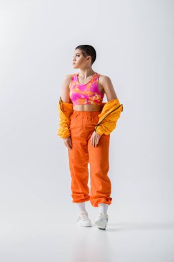 generation z, fashionable young woman with short hair posing in puffer jacket and vibrant pants on grey background, looking away, outerwear, modern subculture, youthful energy, full length 