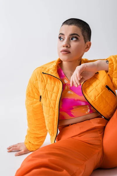 stock image outerwear, fashion statement, tattooed young woman in colorful clothes sitting and looking away on grey background, urban style, individualism, vibrant and youthful energy, stylish look 