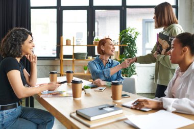 young woman holding magazines and shaking hands with motivation coach while meeting with friendly female community near takeaway drinks in interest club, mutual support and understanding concept clipart