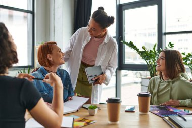 happy african american woman talking to coach near multiethnic members of women interest club in friendly atmosphere with magazines and takeaway drinks, mutual support and understanding concept clipart