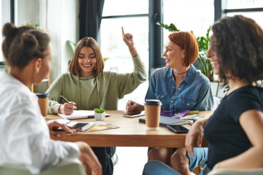 cheerful young woman showing idea gesture and writing in notebook near multiethnic friends and table with paper cups and magazines in female interest club, knowledge-sharing and socializing concept clipart