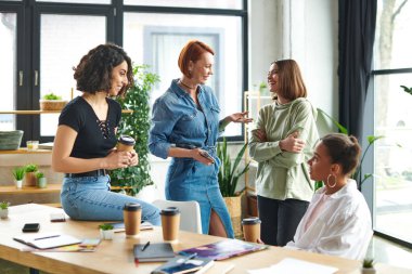 cheerful redhead woman with mobile phone talking to multiracial friend near multiethnic women drinking coffee near magazines and notebooks in interest club, happy leisure of diverse female team clipart