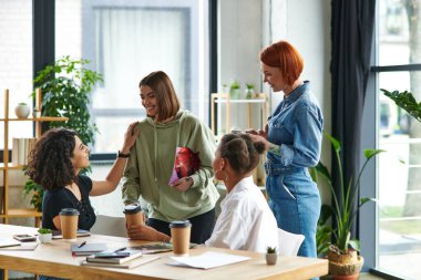 multiracial woman talking to young girlfriend and touching her shoulder near multiethnic members of interest club next to table with magazines and coffee to go, solidarity and understanding concept clipart