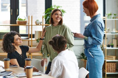 optimistic multicultural female friends talking to pleased young woman during communication near table with paper cups with coffee to go in interest club, solidarity and understanding concept clipart