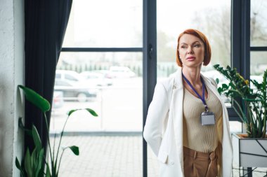 thoughtful psychologist with red hair and name badge standing in white blazer, thinking and looking away in consulting room, professional help and supportive therapy clipart