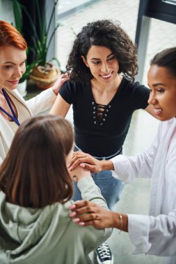 high angle view of diverse group of pleased multicultural women joining hands near positive motivation coach during session in consulting room, moral support and mental wellness concept clipart