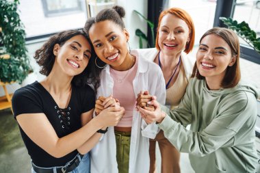 redhead motivation coach and carefree multicultural female friends holding hands and looking at camera during psychology session in consulting room, moral support and mental wellness concept clipart