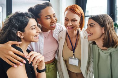 overjoyed redhead psychologist embracing with happy diverse group of multicultural women during motivation session in consulting room, moral support and mental wellness concept clipart