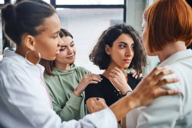 offended multiracial woman looking away near multicultural female friends and psychologist calming her in consulting room, problem-solving and mutual support concept clipart