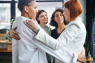 Mutlu ve iyimser çok kültürlü kız arkadaşlar. Kızıl saçlı motivasyon koçuna sarılıyorlar ve danışma odasında destek terapisi, kadın birliği ve destek konsepti sırasında birbirlerine gülümsüyorlar.
