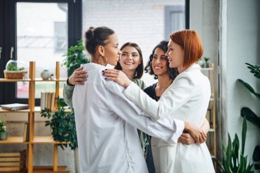 Çok mutlu çoklu etnik kadınlar ve kızıl saçlı motivasyon koçu danışma odasında yan yana dururken sarılıp birbirlerine bakıyorlar, kadın birliği ve destek kavramı.