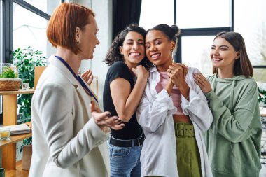 cheerful multiethnic women embracing african american girlfriend near happy motivation coach gesturing in consulting room, female unity and support concept clipart