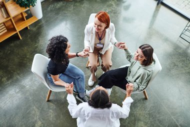 high angle view of joyful multiethnic female friends and redhead motivation coach sitting in circle and holding hands while talking on group therapy, communication and mental wellness concept clipart