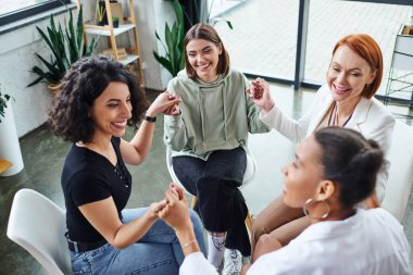 Neşeli çok kültürlü kadın arkadaşlar ve kızıl saçlı motivasyon koçu danışma odasında grup terapisi sırasında el ele tutuşup iletişim kuruyor, arkadaşlık ve zihinsel sağIık kavramı.