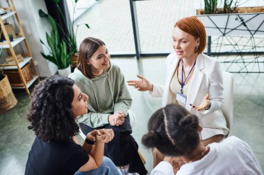 high angle view of redhead psychologist pointing with hands and talking to smiling multicultural women during motivation therapy in consulting room, friendship and mental wellness concept clipart