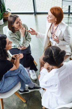 Kızıl saçlı motivasyon koçunun yüksek açılı bakış açısı neşeli genç bir kadını işaret ediyor ve grup terapisi, arkadaşlık ve zihinsel sağIık konsepti sırasında çok ırklı kız arkadaşlarla konuşuyor.