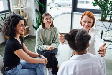 happy redhead motivation coach sitting with outstretched hands and talking to diverse group of multiethnic women during therapy in consulting room, friendship and mental wellness concept clipart