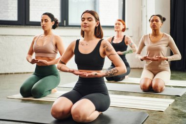 Dövmeli yoga koçu, şimşek altında otururken gözleri kapalı meditasyon yaparken çeşitli etnik gruplara mensup kadınların yanında poz veriyor. İç huzur ve beden farkındalığı kavramı.
