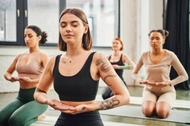 Genç ve dövmeli kadın spor kıyafetleriyle meditasyon yapıyor. Farklı etnik gruplardan oluşan bir grubun yanında bulanık arka plan, iç huzur ve beden farkındalığı kavramı üzerine meditasyon yapıyor.