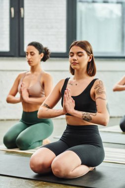 Genç, dövmeli bir kadın şimşek gibi poz veriyor ve kapalı gözlerle meditasyon yapıyor ve çok ırklı bir kız arkadaşın yanında dua eden eller bulanık arka plan, iç huzur ve beden farkındalığı kavramına sahip.
