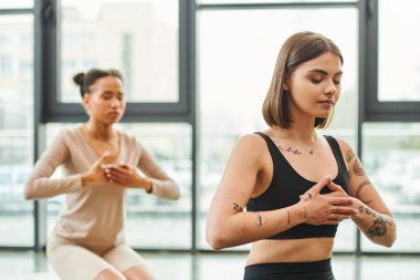 young and tattooed woman meditating with closed eyes and hands on chest during yoga class near african american girlfriend on blurred background, inner peace and body awareness concept clipart