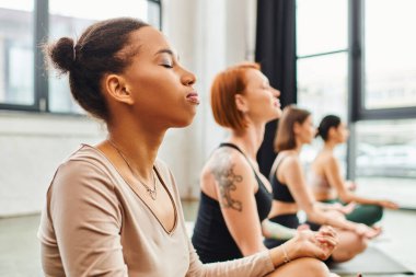 young african american woman meditating with closed eyes near multiethnic friends relaxing in easy pose during yoga class, harmony and wellness concept clipart