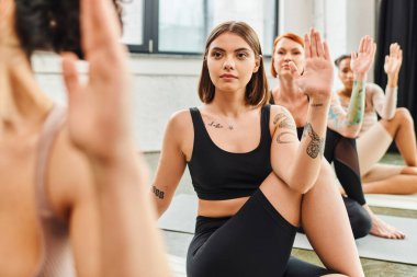 Genç, konsantre ve dövmeli bir kadın spor kıyafetleri içinde balıkların efendisi olarak meditasyon yapıyor yoga dersi sırasında çok ırklı arkadaşlarla, sağlıkla ve zihinsel sağlıkla poz veriyor.