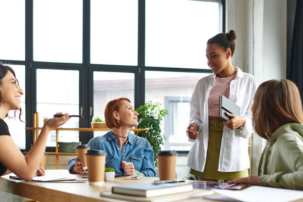 молодая африканская американка, стоящая с ноутбуком и улыбающаяся разнообразной группе мультикультурных подруг возле кофе, чтобы пойти в клуб интересов, взаимной поддержки и понимания концепции