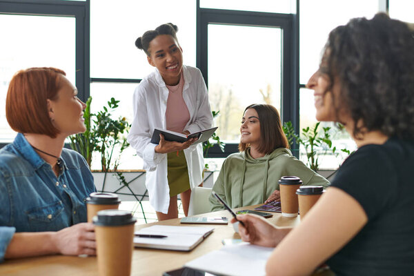 молодая африканская американка с блокнотом, улыбающаяся многонациональным друзьям, сидящим за столом с кофе в дружественной атмосфере интереса клуба, взаимной поддержки и понимания концепции