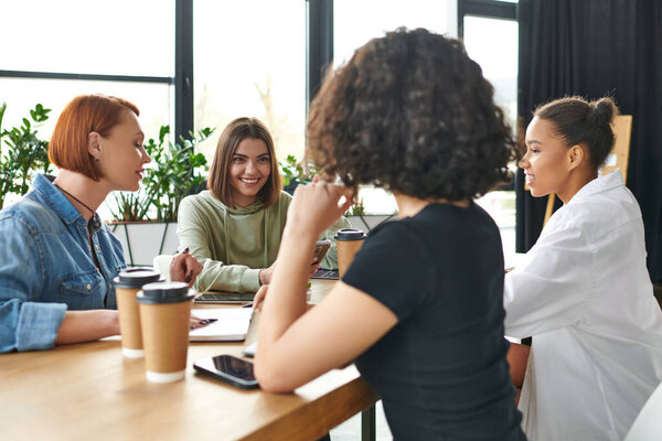 cheerful young woman talking to multiethnic female group near smartphones and coffee to go in friendly atmosphere of interest club, mutual support and understanding concept
