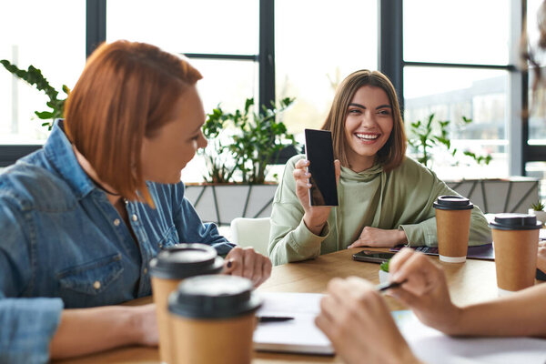 young and optimistic woman showing mobile phone with blank screen to girlfriends spending time with coffee to go in friendly atmosphere of interest club, female friendship and support concept