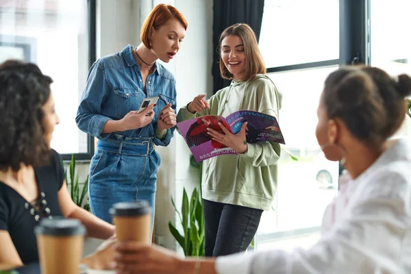 Mujer Sonriente Apuntando Revista Cerca Amigo Sorprendido Con Teléfono Móvil — Foto de Stock