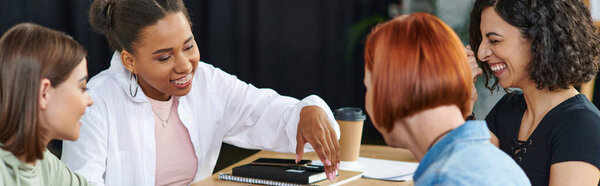 вне себя от радости африканская американская женщина указывая рукой во время общения с мультикультурными подруг возле ноутбуков и вынос напитка в женском клубе, дружественная атмосфера, баннер