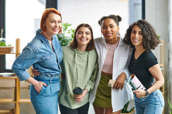 весёлые мультикультурные подруги в стильной повседневной одежде, со смартфоном, бумажной чашкой и журналом, улыбающимися в камеру в женском клубе интересов, взаимной поддержкой, солидарностью и доверием