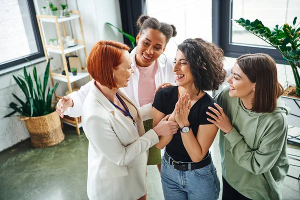 redhead motivation coach and smiling multicultural female friends embracing overjoyed multiracial woman during psychology session in consulting room, moral support and mental wellness concept