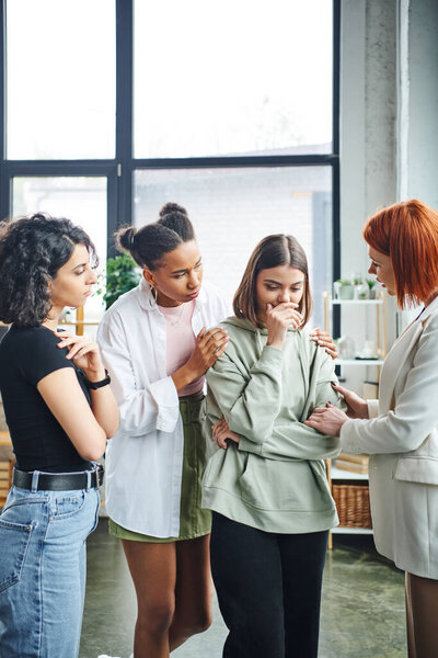 young and depressed woman holding hand near face while standing near psychologist and multicultural female friends in consulting room, problem-solving and psychological help concept