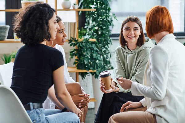 happy young woman sitting with paper cup and listening to redhead psychologist during supportive therapy near multiracial girlfriends, understanding, support and mental health concept
