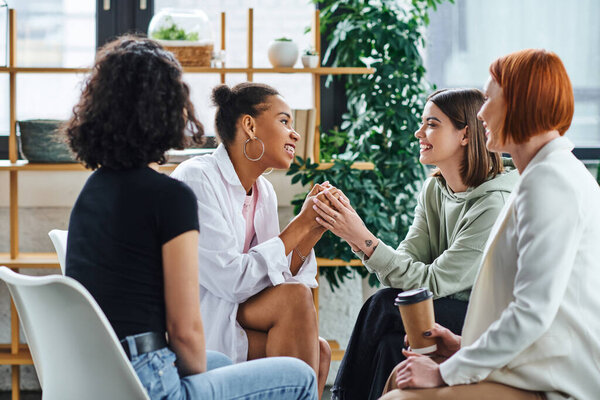 young tattooed woman holding hands of happy african american girlfriend near smiling redhead psychologist sitting with takeaway drink in paper cup, problem-solving and mental wellness concept