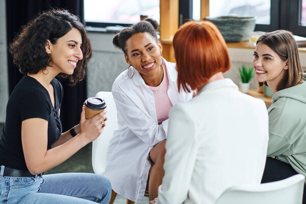 multiracial woman sitting with coffee to go and listening to redhead psychologist together with optimistic multiethnic girlfriends during therapy, understanding, support and mental health concept