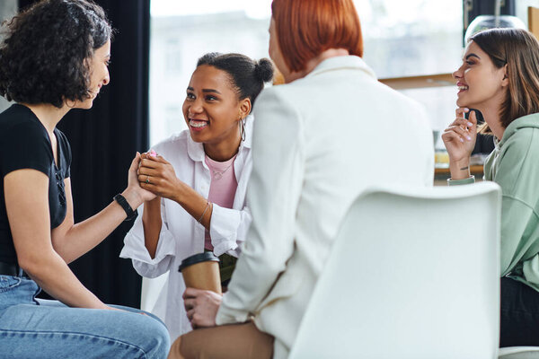 smiling african american woman holding hands with multiracial girlfriend near redhead motivation coach sitting with takeaway coffee on blurred foreground, support and mental health concept