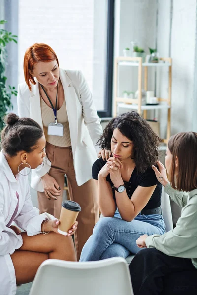 stock image redhead motivation coach and young multiethnic friends calming disappointed multiracial woman during supportive therapy in consulting room, problem-solving and mental wellness concept