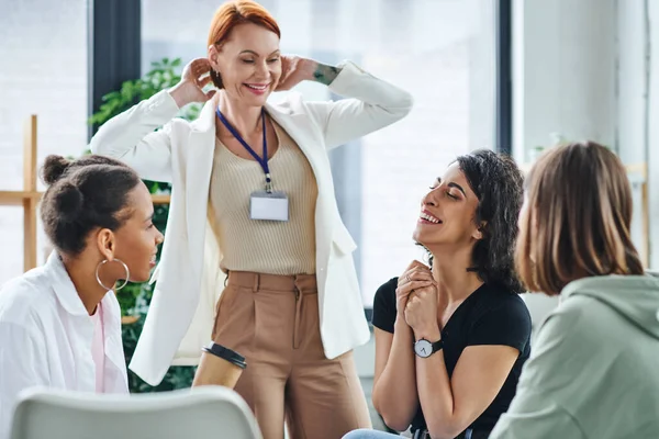pleased motivation coach smiling near diverse multicultural group and multiracial woman laughing with closed eyes during psychology session, understanding, support and mental health concept