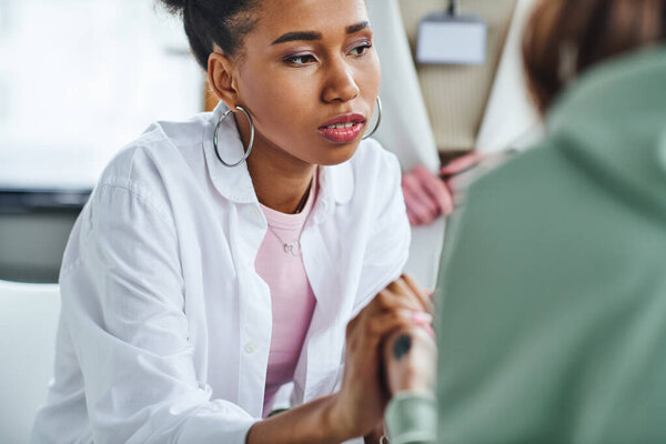 young and serious african american woman holding hands of friend near professional psychologist on blurred background in consulting room, empathy, solidarity and problem-solving concept