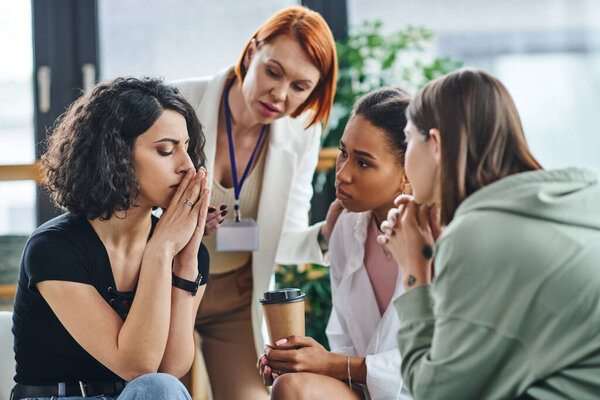 professional psychologist and young multiethnic girlfriends looking at depressed multiracial woman sitting with closed eyes and hands near face in consulting room, problem-solving concept