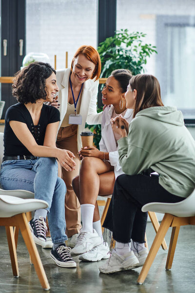 happy multiracial woman sitting near smiling multiethnic female friends and redhead motivation coach on supportive group therapy in consulting room, friendship and mental wellness concept