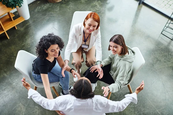 high angle view of positive multicultural women and redhead psychologist gesturing while sitting in circle on motivation group therapy in consulting room, communication and mental wellness concept