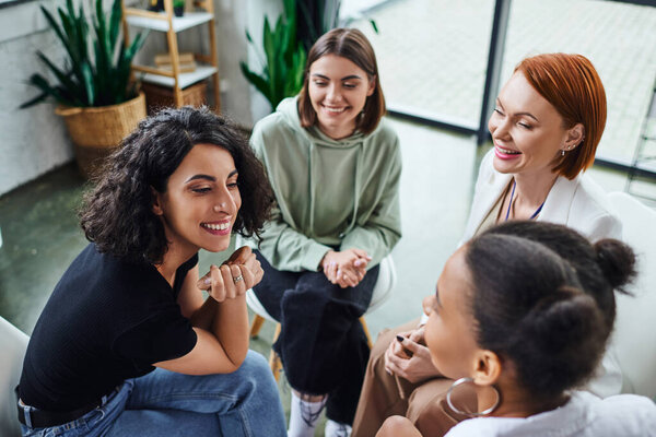 high angle view of cheerful multiracial woman talking to african american girlfriend near smiling psychologist during group therapy in consulting room, communication and mental wellness concept